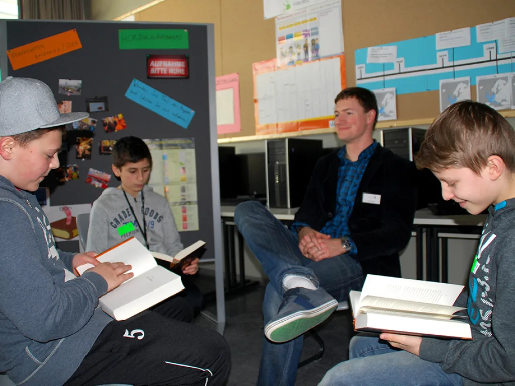 Zwei Jugendliche und ein Erwachsener sitzen mit Büchern im Gespräch.